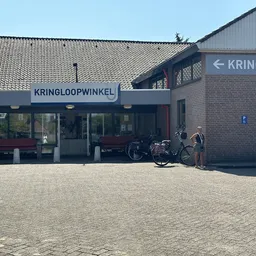 Voorgevel van kringloopwinkel met blauwe luifel en winkelnaam, fiets geparkeerd ervoor