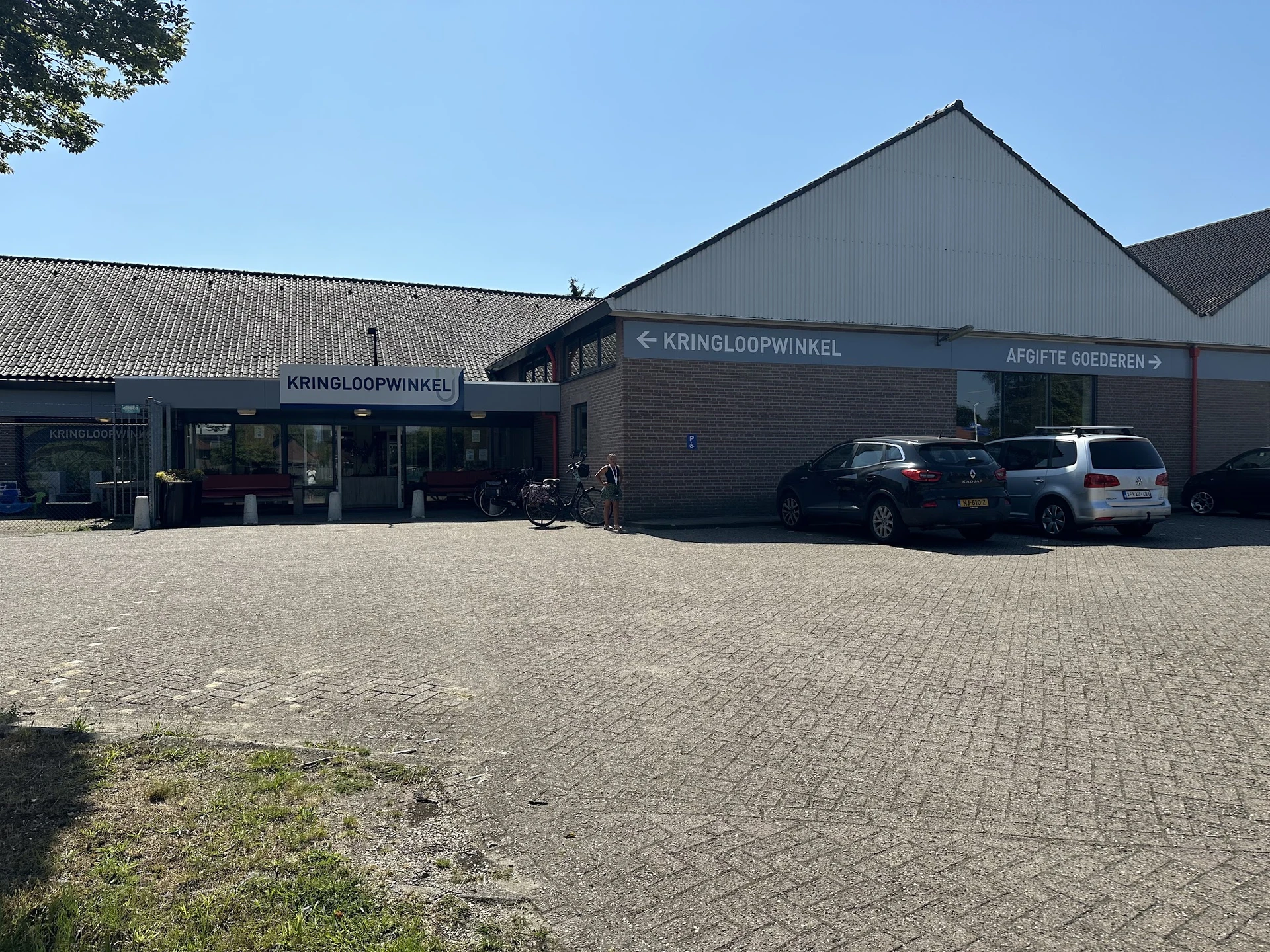 Voorgevel van kringloopwinkel Verderest met parkeerplaats en twee auto's, heldere dag