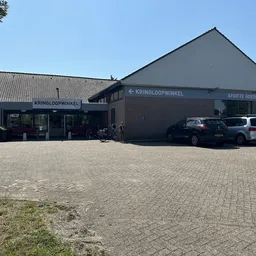 Voorgevel van kringloopwinkel Verderest met parkeerplaats en twee auto's, heldere dag