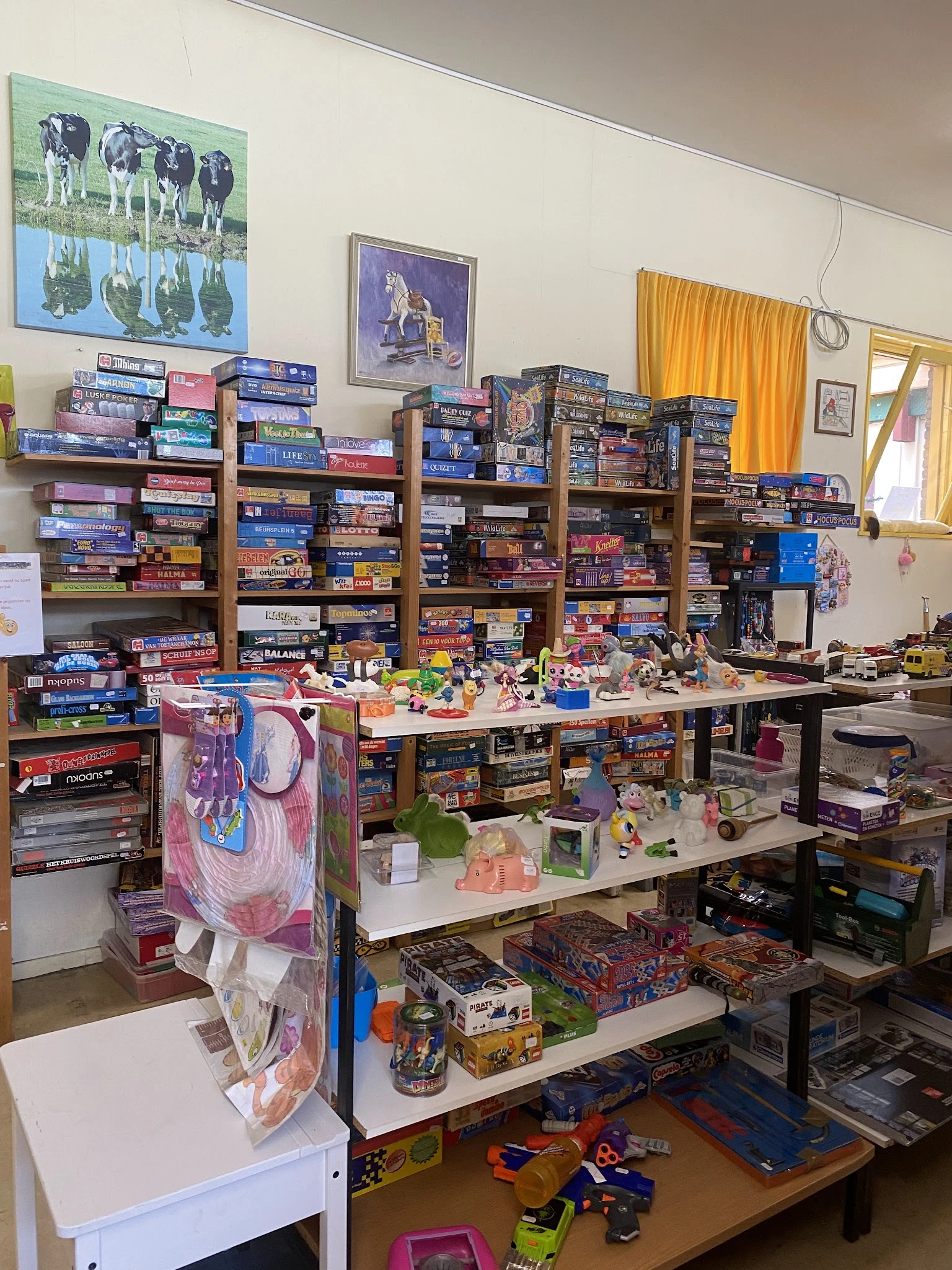 Winkelvloer met houten stellingen vol dozen bordspellen, tafels met speelgoed en verzamelaarsartikelen, gele gordijnen en pos