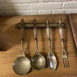 Vier vintage keukengereedschappen met houten handvaten aan een muurmontage: vergiet, soeplepel, schuimspaan en vork.