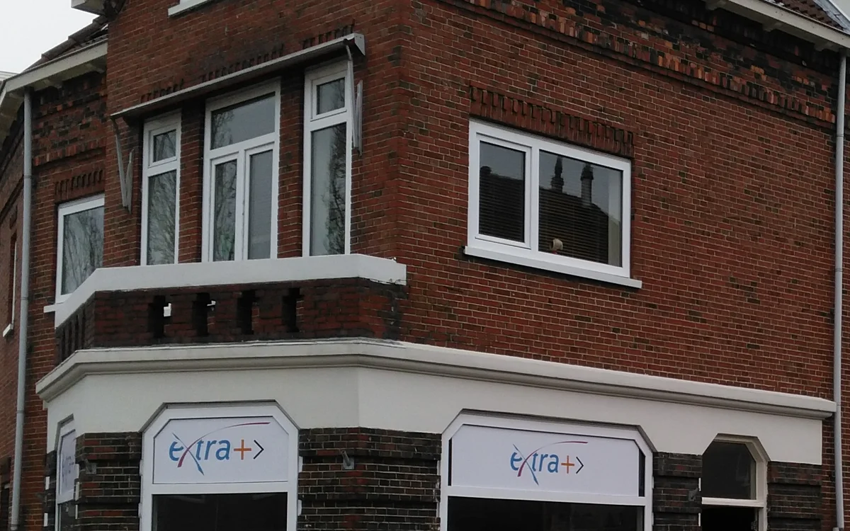 Gevel van kringloopwinkel ExtraPlus met rode bakstenen muur, witte raamkozijnen en winkelruit met logo op begane grond