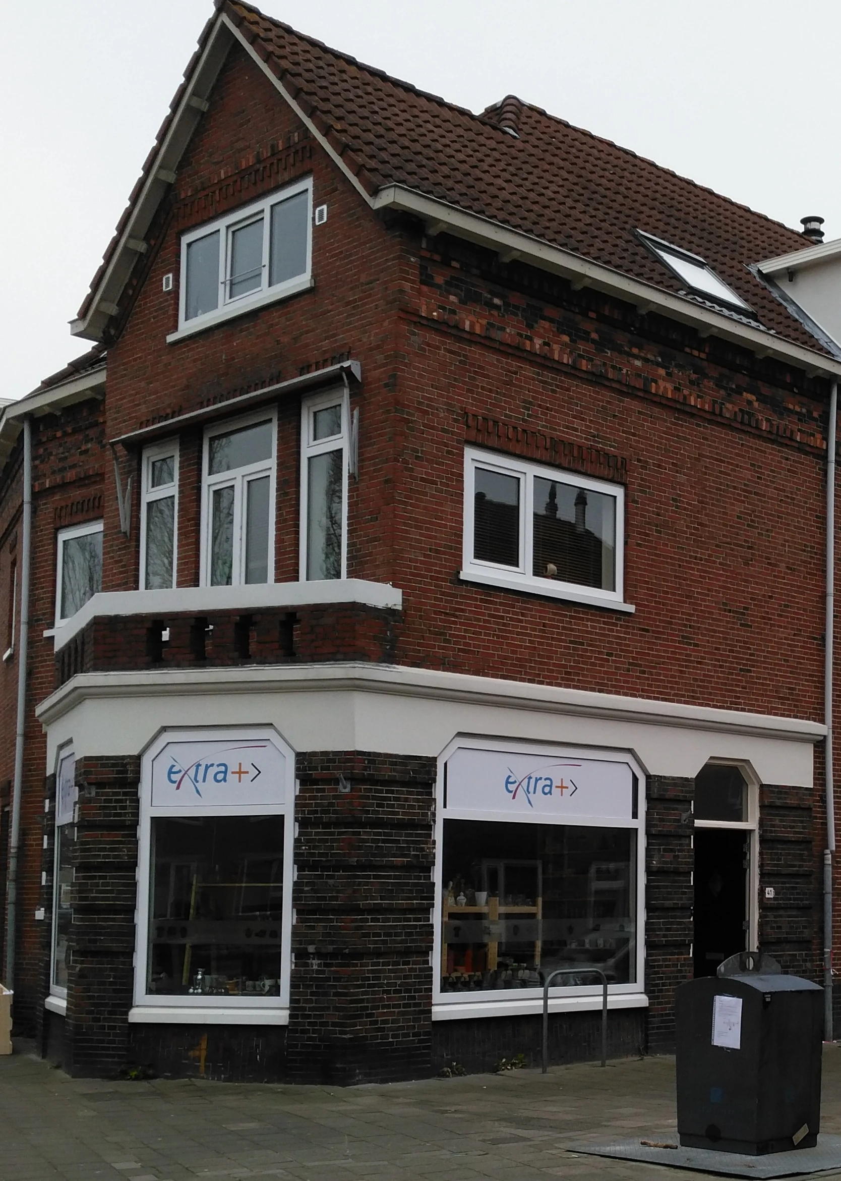 Gevel van kringloopwinkel ExtraPlus met rode bakstenen muur, witte raamkozijnen en winkelruit met logo op begane grond