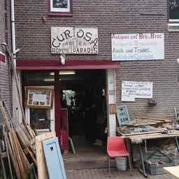 Voorkant van kringloopwinkel met rode deur, uitstallingen buiten op tafel en tegen de gevel, borden met teksten
