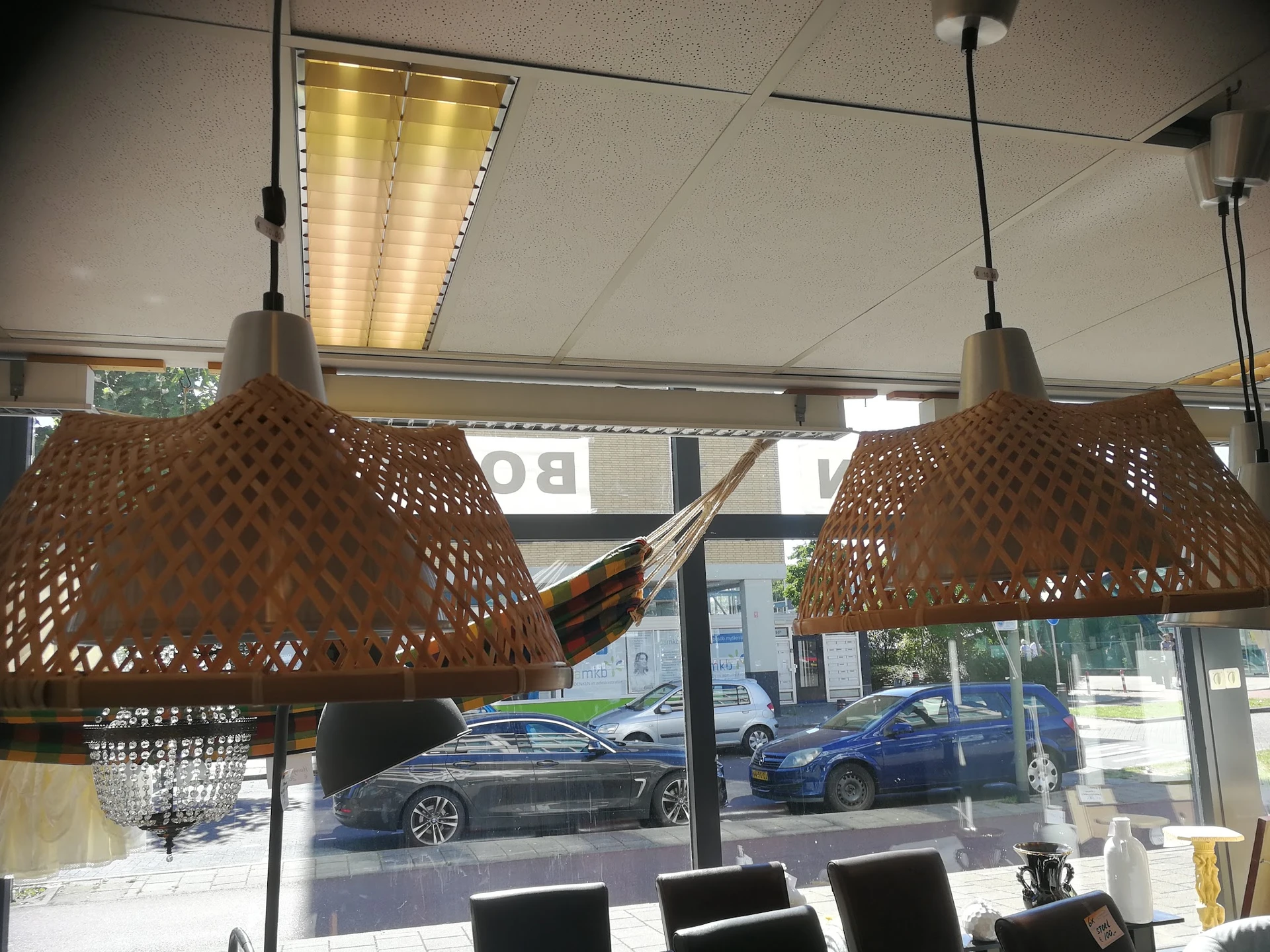 Interieur van kringloopwinkel met twee bruine hangende lampen boven zwarte stoelen, uitzicht op straat met geparkeerde auto's