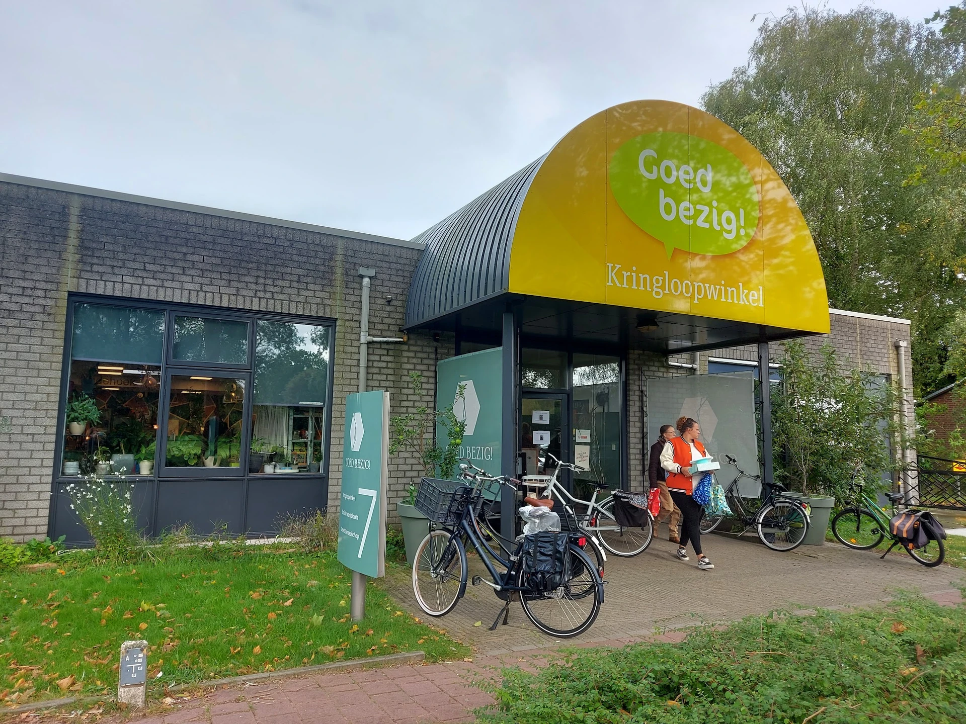 Voorkant van kringloopwinkel met gele luifel, bakstenen gevel en fietsen ervoor geparkeerd