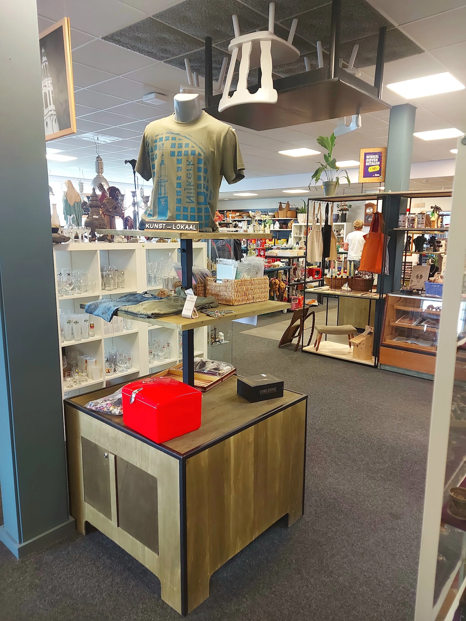 Winkelvloer met stellingen vol spullen, kleding aan hangers en een rode doos op een tafel