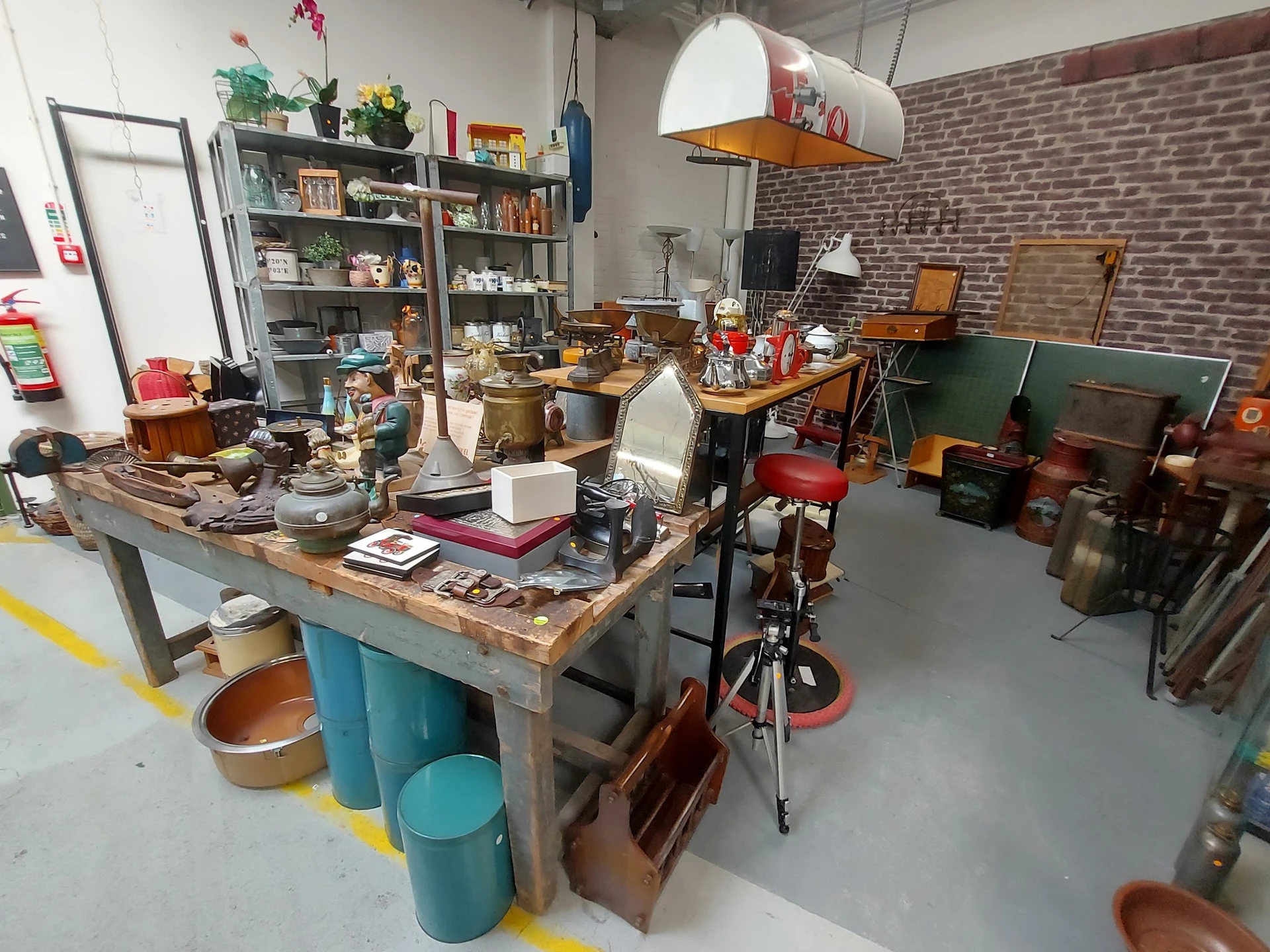 Werkplaats of opslagruimte met een centrale tafel vol spullen, stellingen met boeken en huishoudartikelen, bakken en diverse 