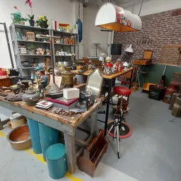 Werkplaats of opslagruimte met een centrale tafel vol spullen, stellingen met boeken en huishoudartikelen, bakken en diverse 