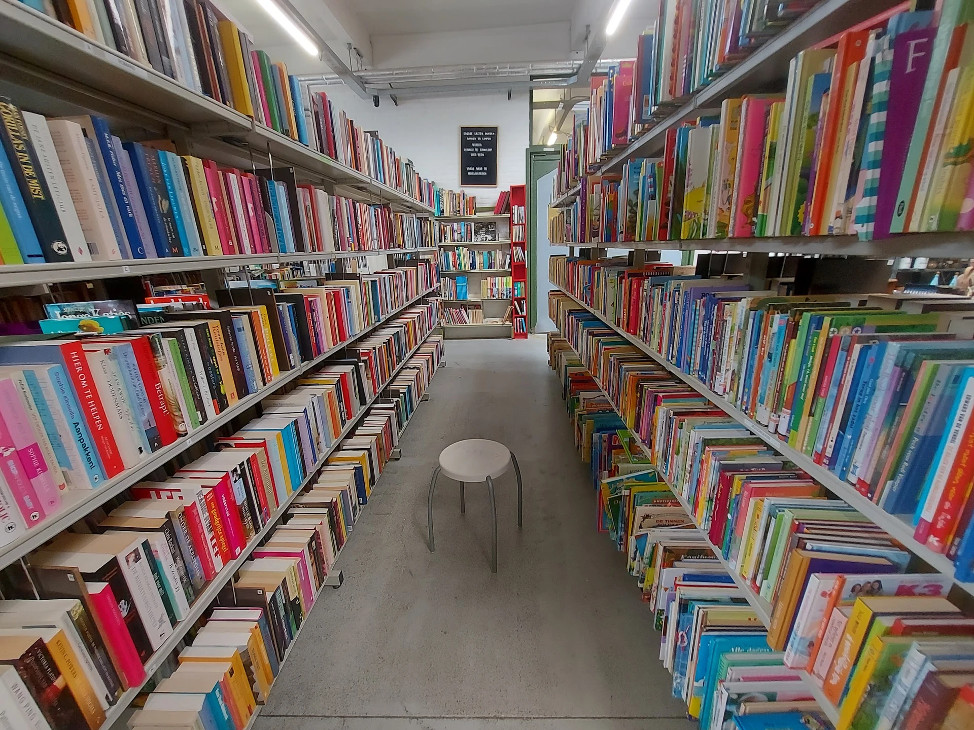 Boekenrekken in een kringloopwinkel met gekleurde boegenrijen aan beide zijden van een gangpad, met een witte kruk in het mid
