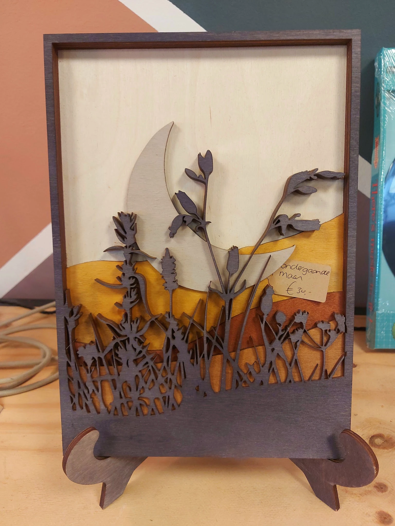 Decoratief kunstwerk van donkerpaars metaal met bloemen- en grassen-silhouetten tegen geel en oranje achtergrond