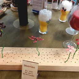 Houten bloemschikbord met bloemen en prijskaartje, omgeven door glazen met dranken op een grijze tafel