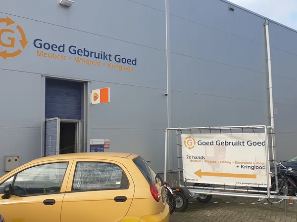 Grijze gevel van kringloopwinkel met oranje logo en naam 'Goed Gebruikt Goed', gele auto op parkeerplaats