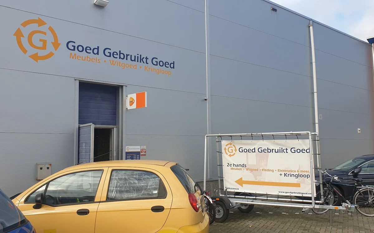 Grijze gevel van kringloopwinkel met oranje logo en naam 'Goed Gebruikt Goed', gele auto op parkeerplaats
