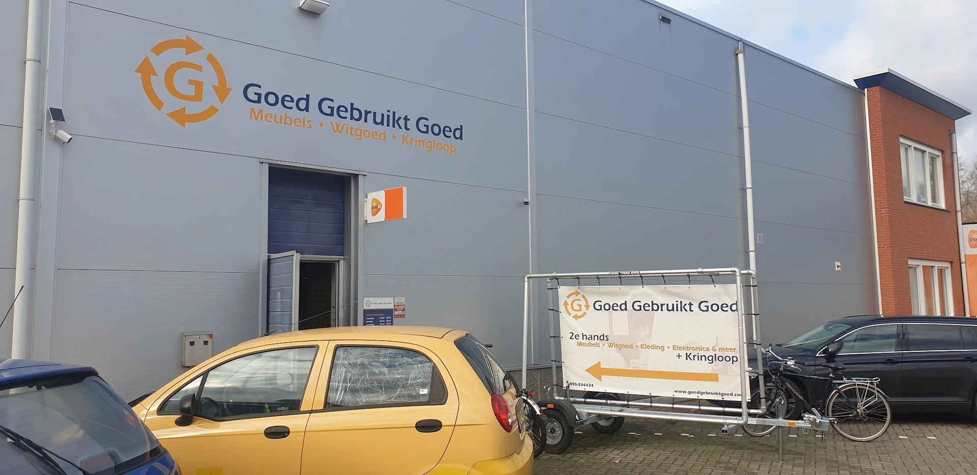 Grijze gevel van kringloopwinkel met oranje logo en naam 'Goed Gebruikt Goed', gele auto op parkeerplaats
