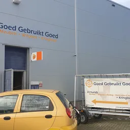 Grijze gevel van kringloopwinkel met oranje logo en naam 'Goed Gebruikt Goed', gele auto op parkeerplaats
