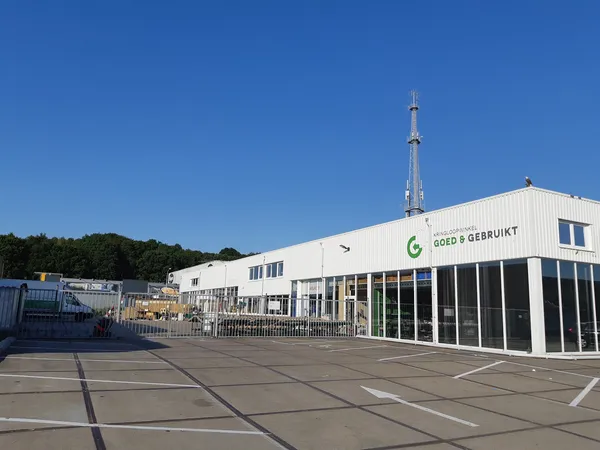 Witte gevel van kringloopwinkel Goed & Gebruikt met grote ramen en groene logo, parkeerterrein ervoor