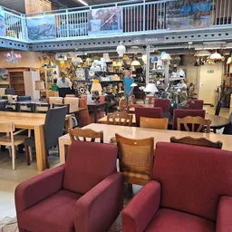 Winkelvloer met bordeauxrode fauteuils, houten tafels en stoelen, daarachter meubels en huishoudartikelen