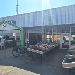 Voorkant van kringloopwinkel met groene houten afdak, gesloten deuren en uitstallingen op het terras
