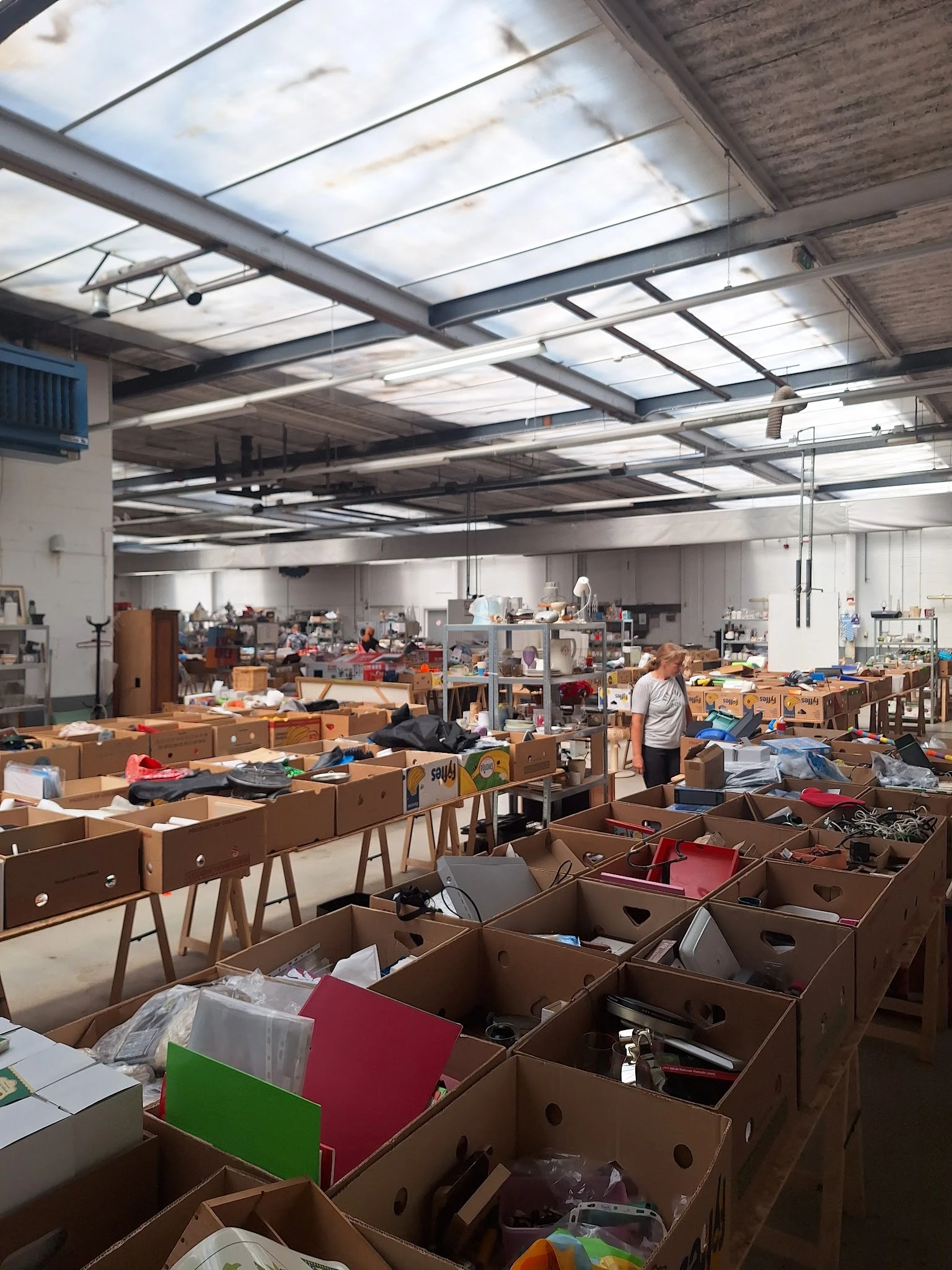 Ruim magazijn met houten meubels, dozen en spullen verspreid over tafels en op de vloer, enkele medewerkers aan het werk