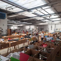 Ruim magazijn met houten meubels, dozen en spullen verspreid over tafels en op de vloer, enkele medewerkers aan het werk
