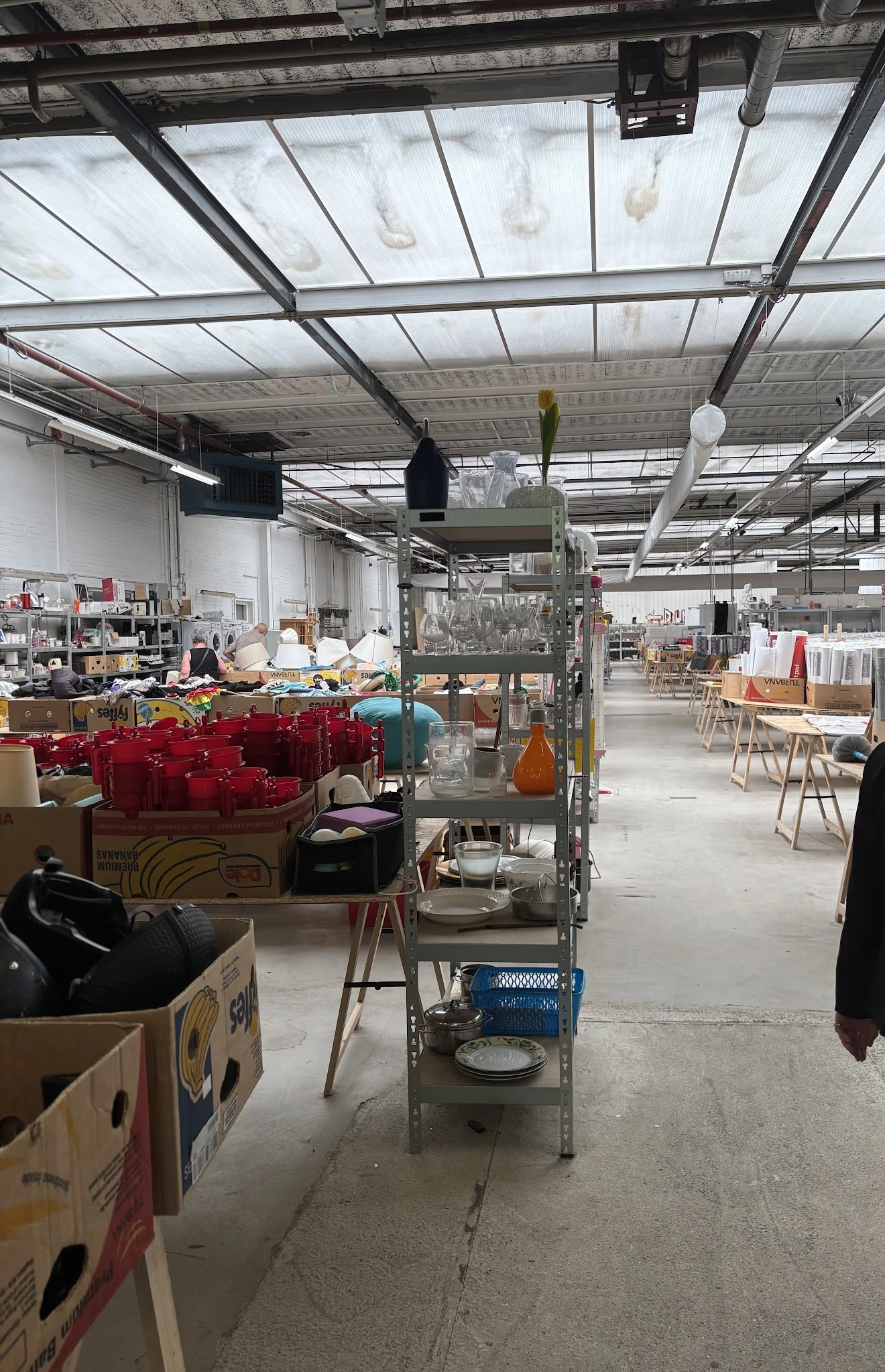Ruim magazijn van kringloopwinkel met stellingen, dozen met rode spullen, meubilair en diverse artikelen verspreid over de vl