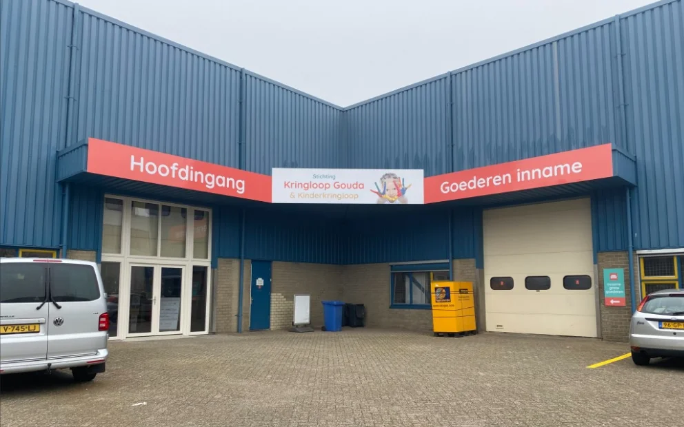 Voorkant van Kringloop Gouda met blauwe golfplaten gevel, rode banner met winkelnaam, glazen deuren en gele brievenbus