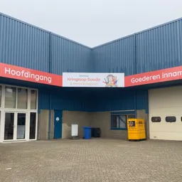 Voorkant van Kringloop Gouda met blauwe golfplaten gevel, rode banner met winkelnaam, glazen deuren en gele brievenbus