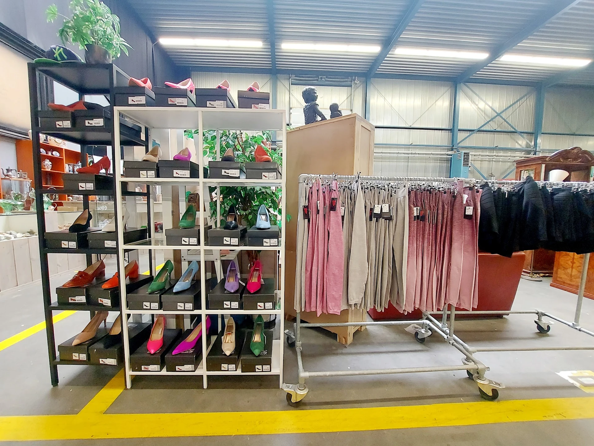 Uitgestalde kledingrekken met roze, beige en zwarte jurken naast een zwarte stellingkast met schoenen en accessoires