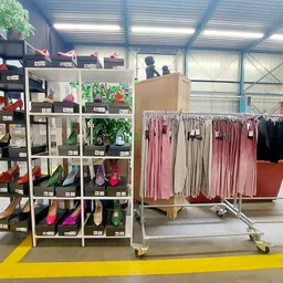 Uitgestalde kledingrekken met roze, beige en zwarte jurken naast een zwarte stellingkast met schoenen en accessoires