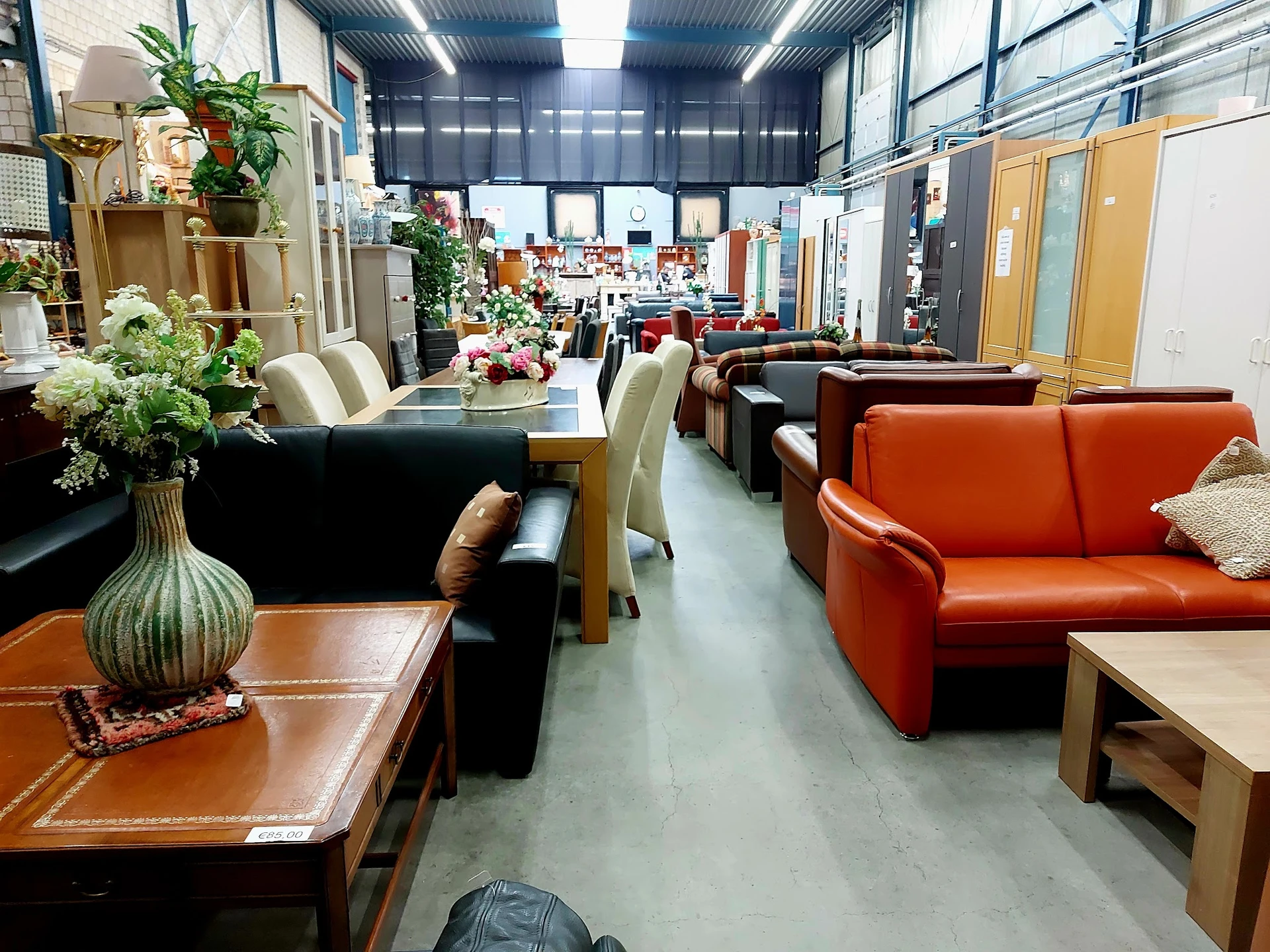 Winkelvloer met diverse meubels: zwarte tafel met stoelen, oranje bank, houten tafels en plantendecoratie