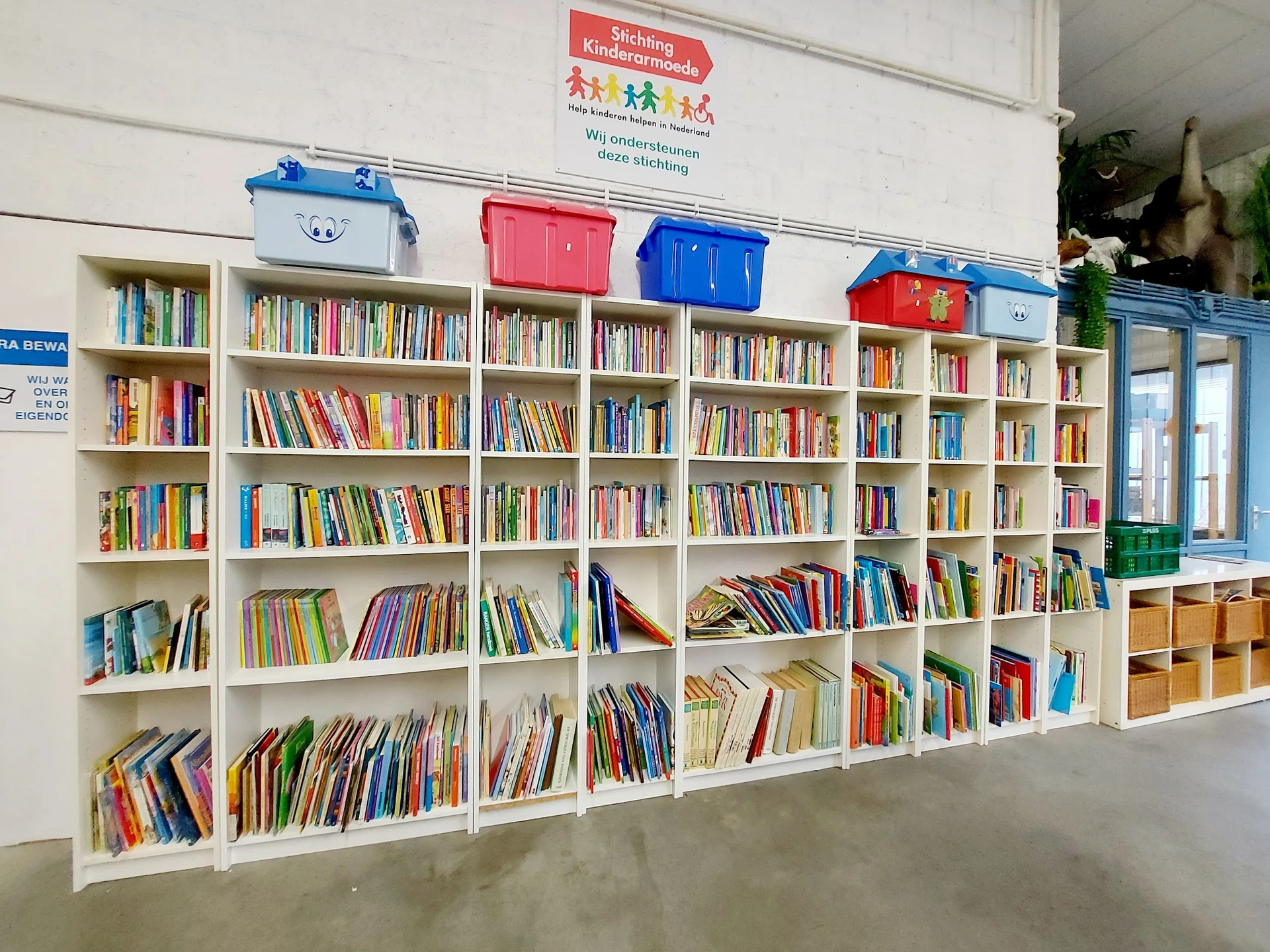Witte boekenkasten vol met tweedehands boeken in verschillende kleuren, met gekleurde opbergboxen bovenop