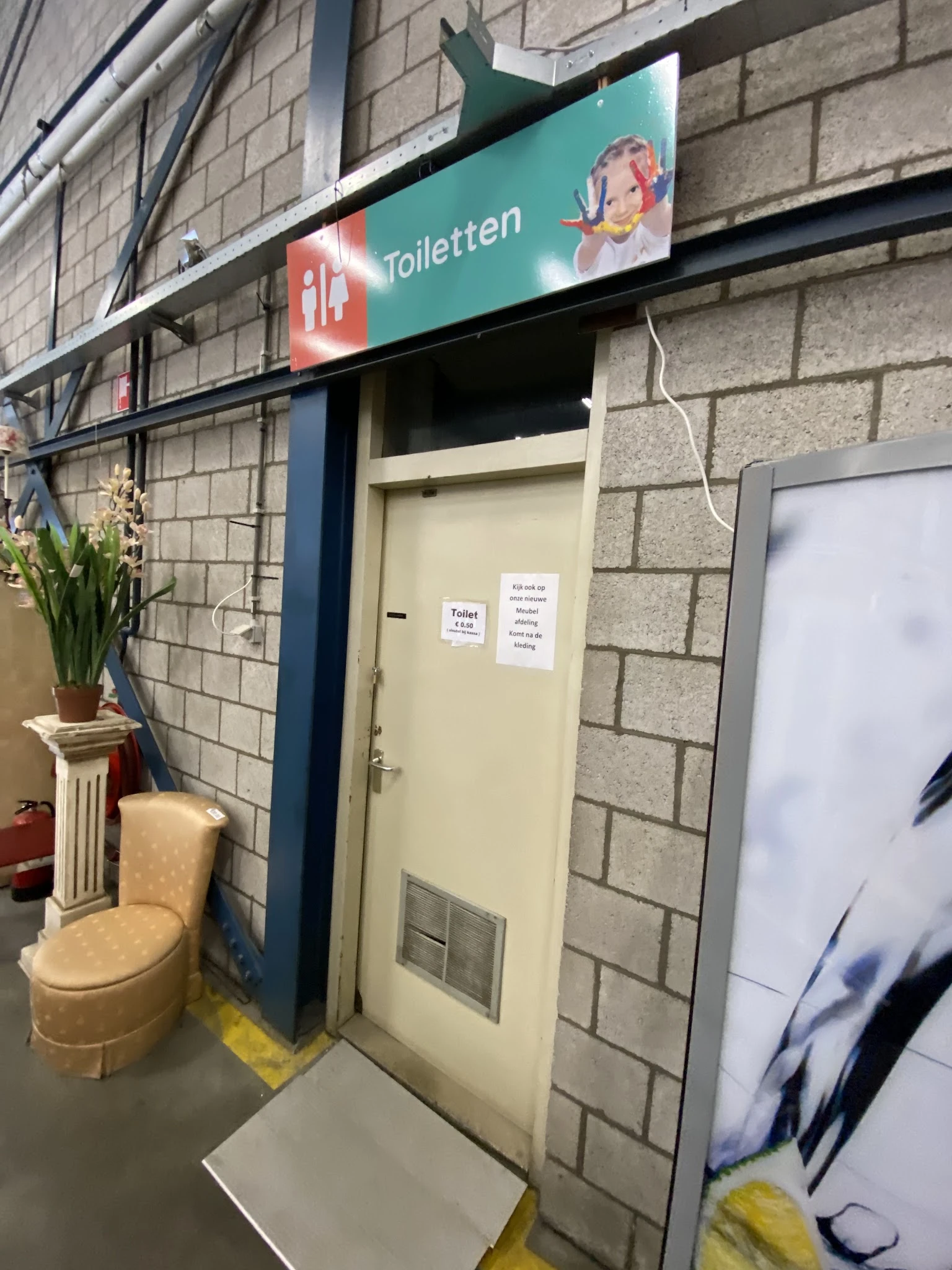 Ingang van kringloopwinkel met gele deur, bloempot en exit-bord zichtbaar