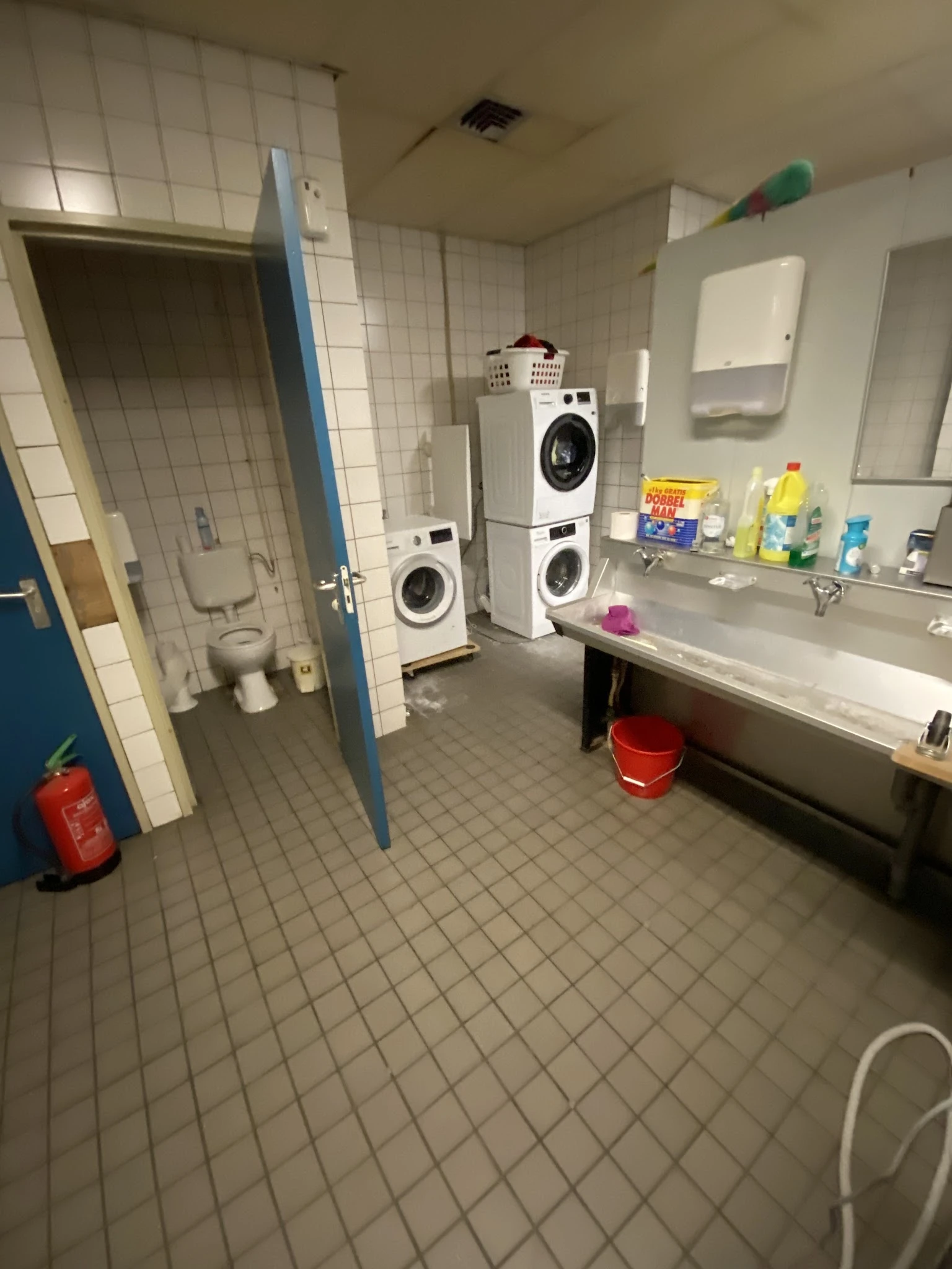 Badkamer met wasmachines, wastafel, toiletruit en schoonmaakspullen op planken