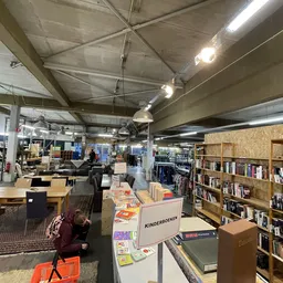 Ruim winkelbinnenland met boekenkasten, tafels met spullen, en hoog plafond met industriële verlichting