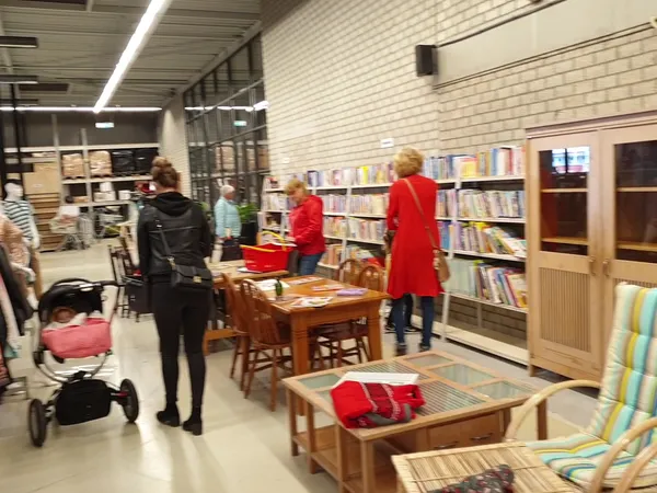 Binnenaanzicht van kringloopwinkel met boekenrekken, houten tafels, zithoeken en bezoekers die rondkijken