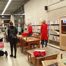 Binnenaanzicht van kringloopwinkel met boekenrekken, houten tafels, zithoeken en bezoekers die rondkijken