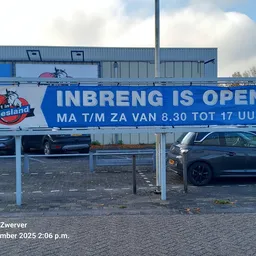 Blauwe openingsbanner 'INBRENG IS OPEN' boven ingang kringloopwinkel met parkeerplaats en geparkeerde auto's