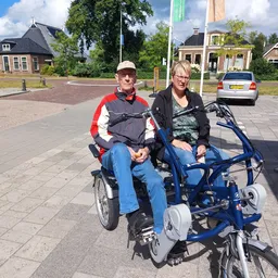 Twee personen op een blauwe driewieler op een straatje met huizen op de achtergrond