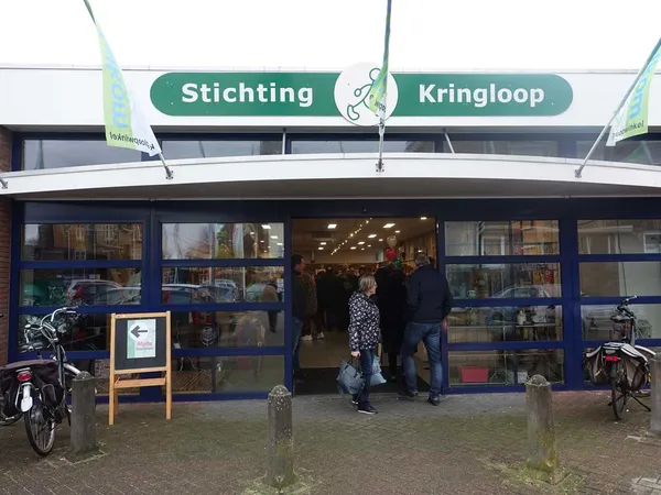 Gevel van kringloopwinkel met groene luifel, naambordje 'Stichting Kringloop', glazen deuren en ramen, enkele bezoekers bij i