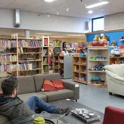 Ruime winkelvloer met boekenkasten, zithoeken met grijze en witte fauteuils, rode kussens, en verschillende kasten met spulle