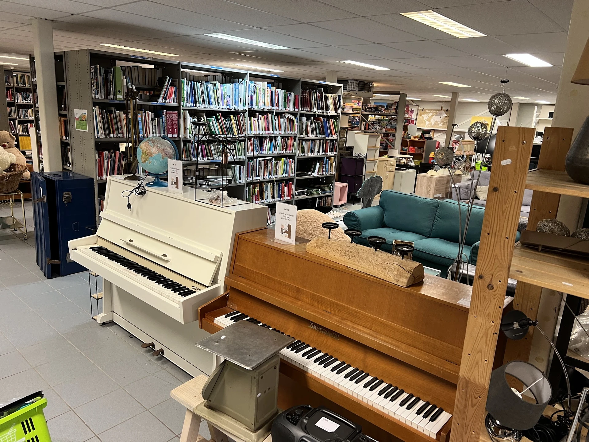 Binnenaanzicht van kringloopwinkel met pianino's, boekenkasten, zithoek en diverse spullen