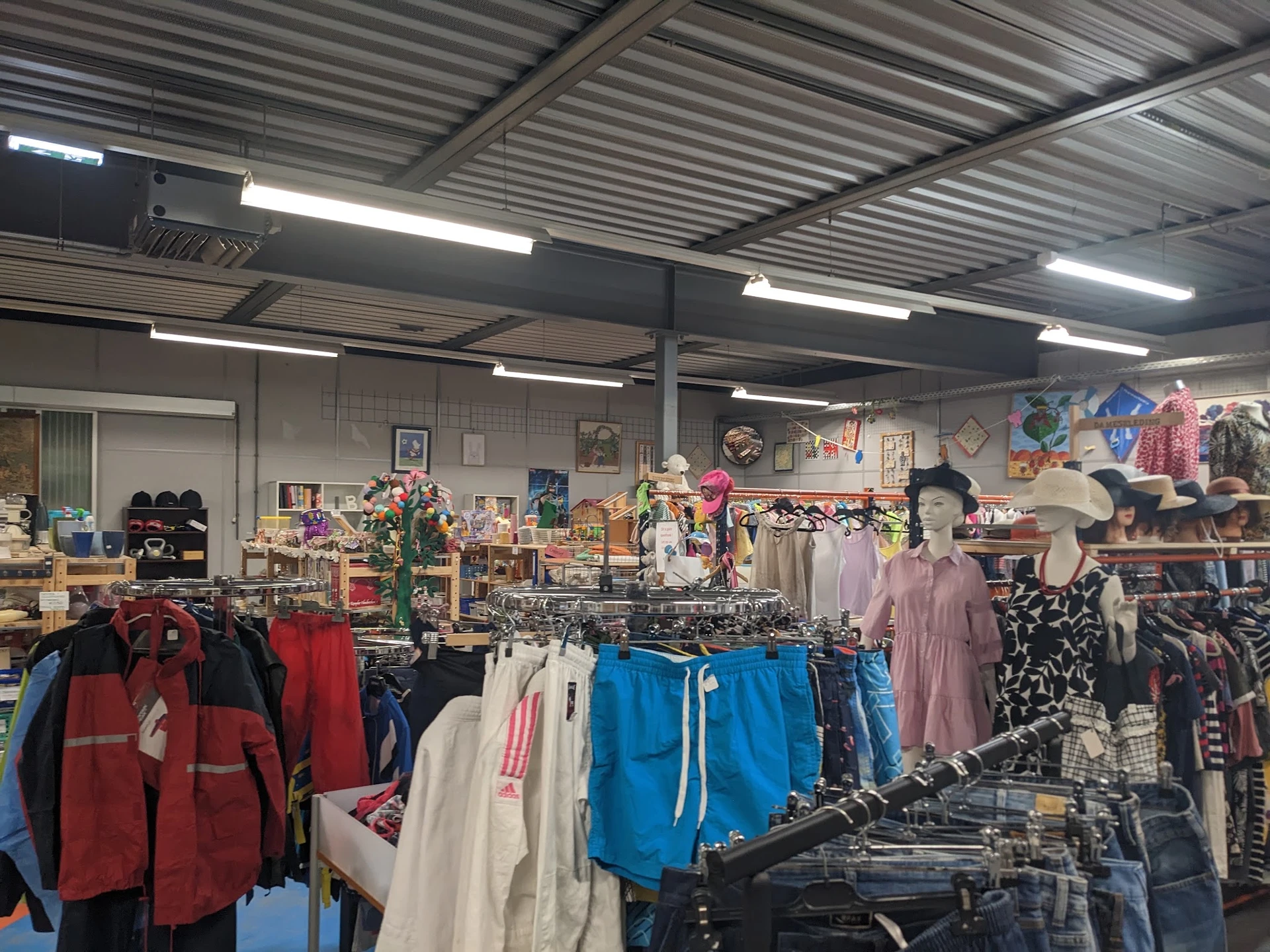 Winkelvloer met rekken vol tweedehands kleding, verlicht met TL-buizen, achtergrond met decoratie en verdere artikelen