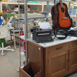 Houten dressoir met witte marmeren blad, met daarop een typemachine, gitaar en andere spullen in een kringloopwinkel