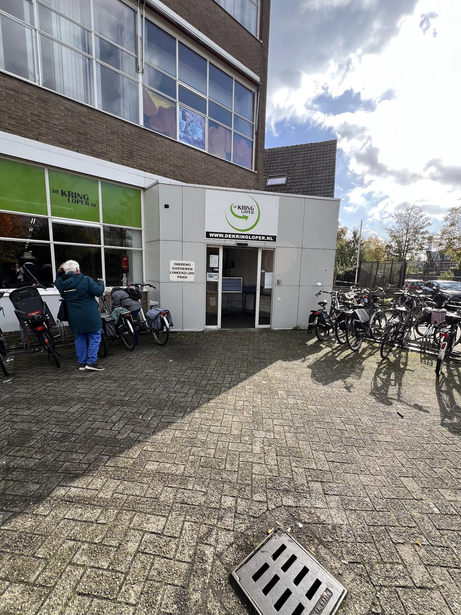Witte gevel van kringloopwinkel met groene zijpui, ingang met glazen deuren, fietsen geparkeerd ervoor
