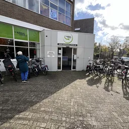 Witte gevel van kringloopwinkel met groene zijpui, ingang met glazen deuren, fietsen geparkeerd ervoor