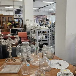 Glazen vazen en kannen op houten tafel in kringloopwinkel, op achtergrond interieur met meubels en stellingen