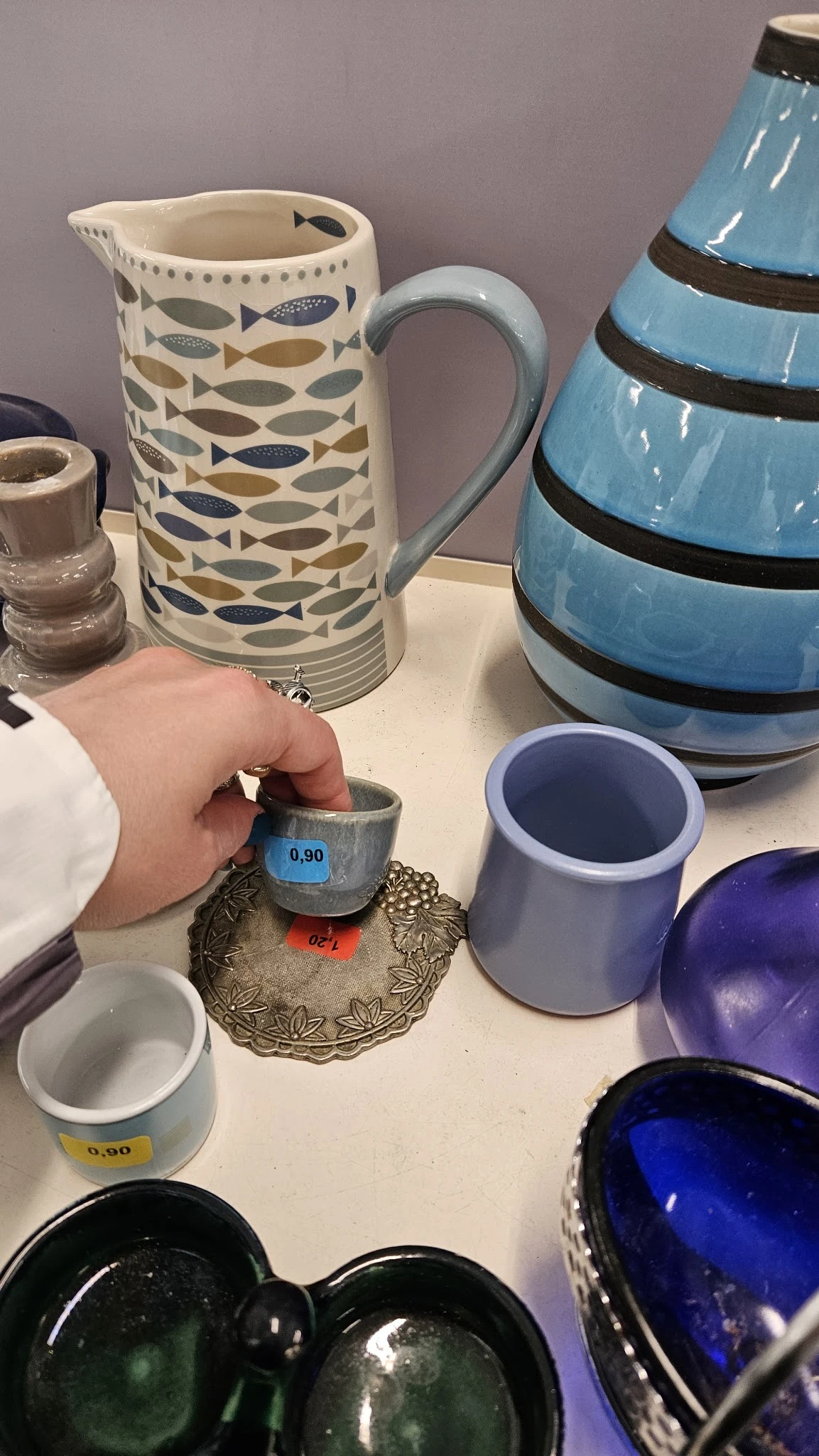 Hand houdt grijze kommetje met prijssticker vast, omgeven door ander servies en huishoudartikelen op tafel