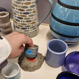 Hand houdt grijze kommetje met prijssticker vast, omgeven door ander servies en huishoudartikelen op tafel