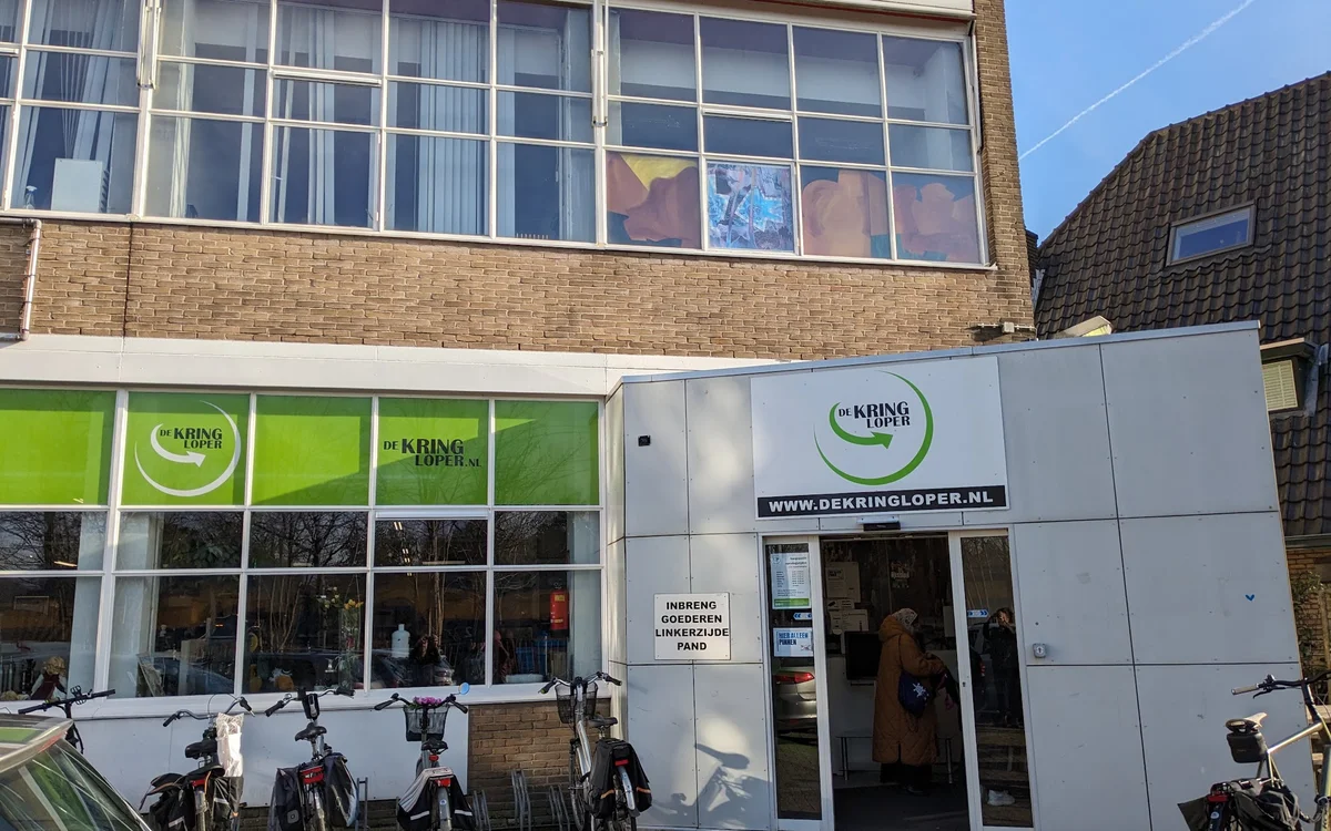 Witte gevel van kringloopwinkel met groene ramen en ingang, fietsen buiten geparkeerd